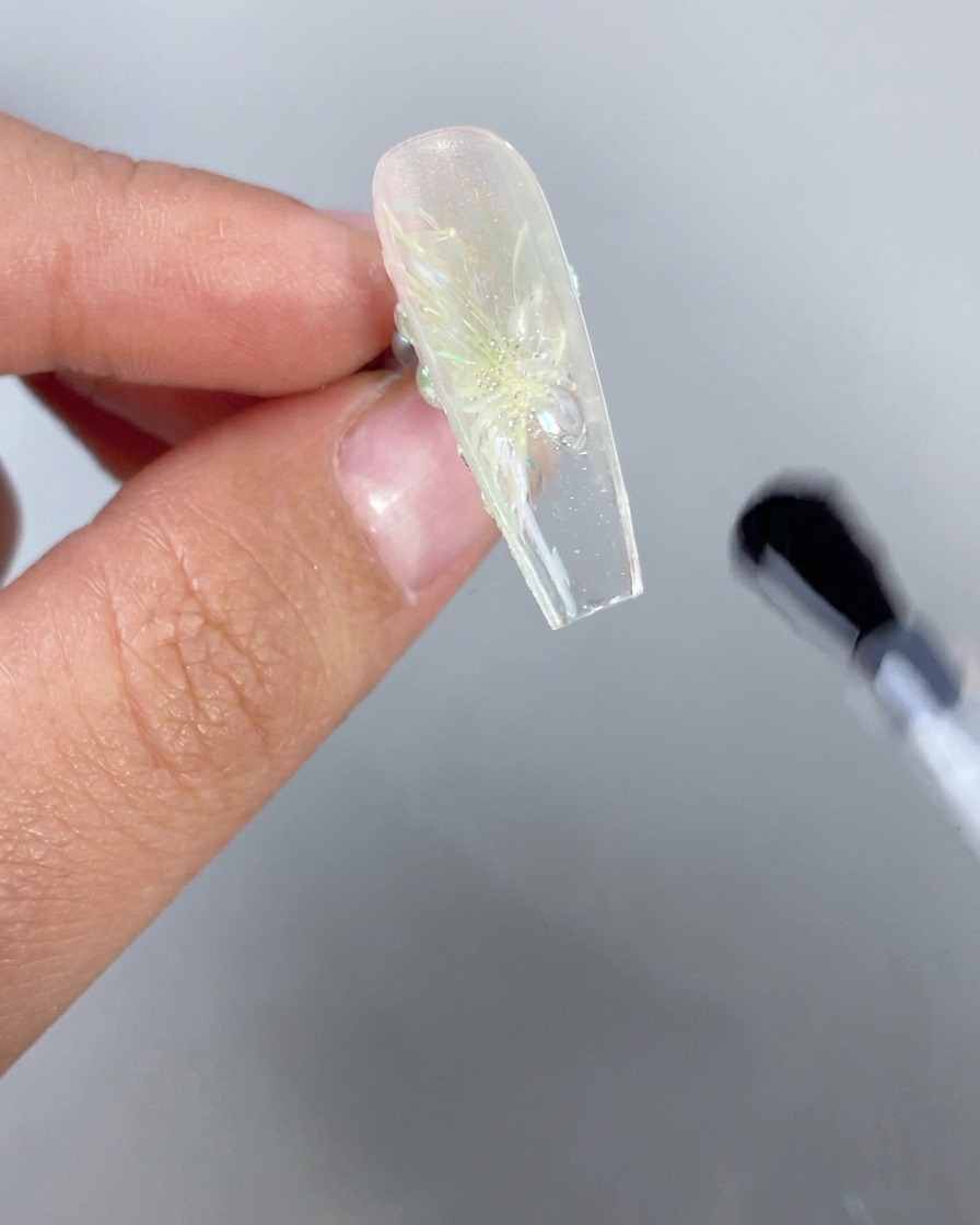 Eine Nahaufnahme einer Hand, die einen eis-transparenten Nagel mit einem 3D-Blumendesign verkehrt herum hält, während ein Pinsel klaren Überzug auf die Unterseite der freien Kante aufträgt.
