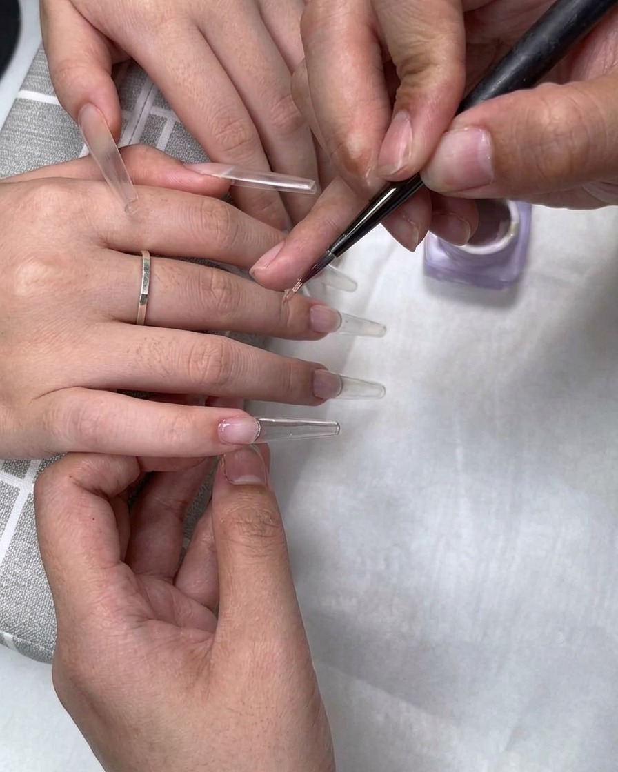 Ein feiner Pinsel trägt Milchkaffee-Nude-Gel-Politur nahe der Nagelhaut einer klaren Nagelverlängerung auf.