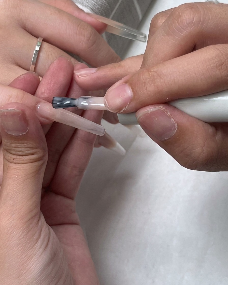 Ein Pinsel trägt eine Schicht transparenten Nude-Lacks über die gesamte Nageloberfläche auf.