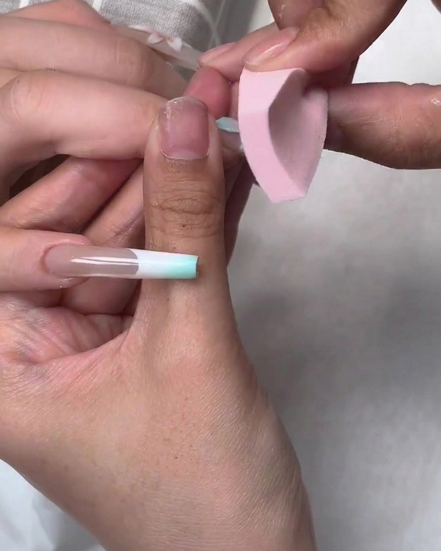 Ein kleiner rosa Schwamm tupft blaues Gel-Polish auf die Ränder einer Nagelapplikation während eines Nail-Art-Tutorials.