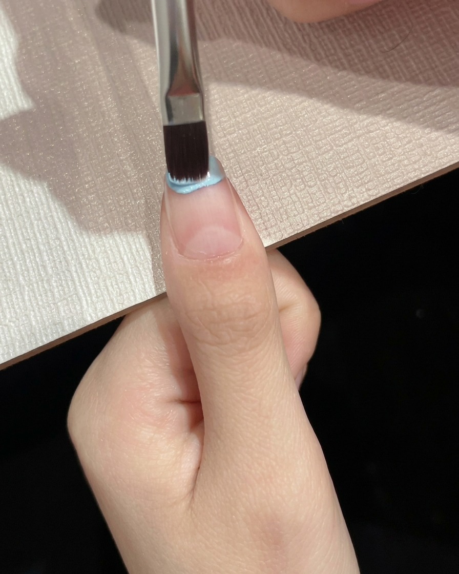 Ein Flachpinsel trägt hellblauen Gelack auf die obere Hälfte einer Nagelplatte auf.