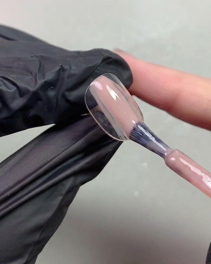 Eine Nahaufnahme einer schwarz bemalten Hand, die mit einem Pinsel transparentes, nudefarbenes Gel auf eine Nagelspitze aufträgt.