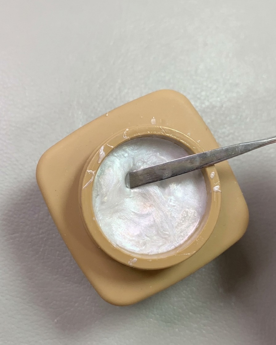Ein Spatel schöpft schimmerndes perlmuttartiges Modelliergel aus einem kleinen runden Tiegel.
