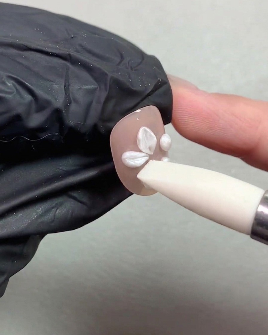 Ein Silikonpinsel drückt und formt weiße Perl-Gel-Kugeln zu Blütenblättern auf einem Nagel.