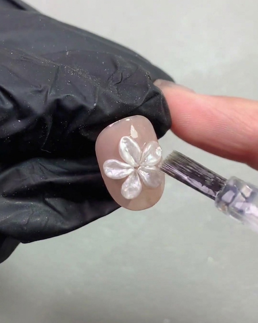 Ein Nagelpinsel trägt vorsichtig eine dünne Topcoat-Schicht um eine Perlenblume auf.