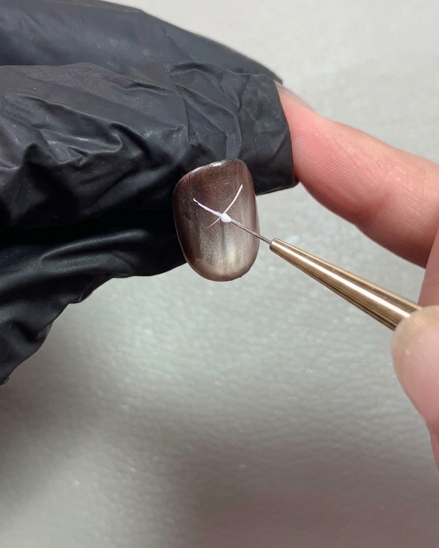 Ein feiner Liner-Pinsel zeichnet dünne weiße Linien, um eine Schleife auf einem dunklen Nagel zu formen.
