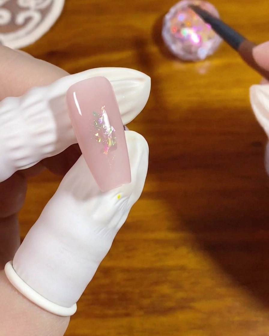 Ein flacher Gelpinsel drückt kleine irisierende Opalflocken auf die Mitte eines rosa lackierten Nagels.
