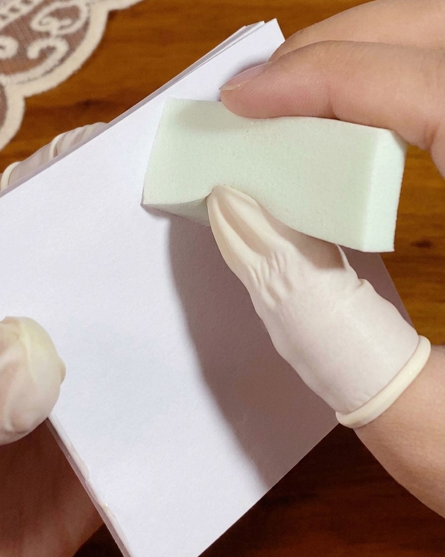 Eine behandschuhte Hand drückt einen mit weißem Gel beladenen Schwamm vor dem Auftragen auf die Nägel auf ein Palettenpapier.