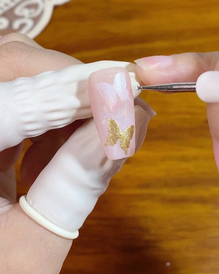 Ein feiner Detailpinsel trägt winzige weiße Punkte neben einem goldenen Schmetterlings-Nagelelement auf.
