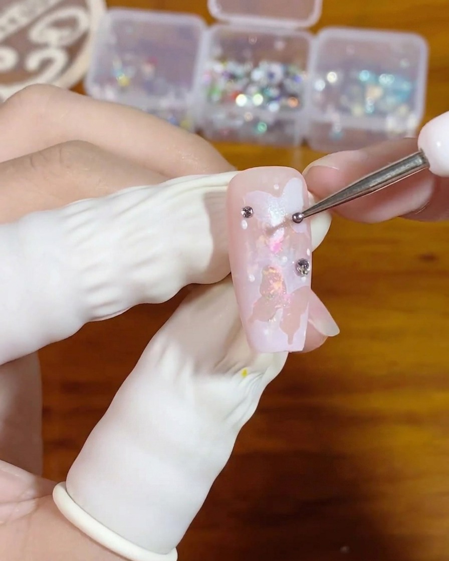 Eine Nahaufnahme einer Hand, die mit einem feinen Dotting-Tool ein winziges Tröpfchen Klargel auf einen rosa Nagel mit weißer Schmetterlings-Nail-Art aufträgt.