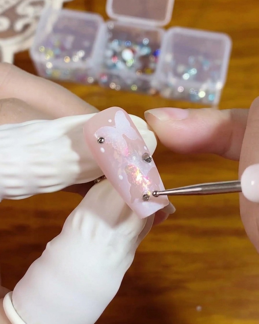 Ein Dotting-Tool platziert einen kleinen silbernen Flatback-Strassstein auf einer Klargelperle auf einem rosa Nagel mit Schmetterlingsdesign.