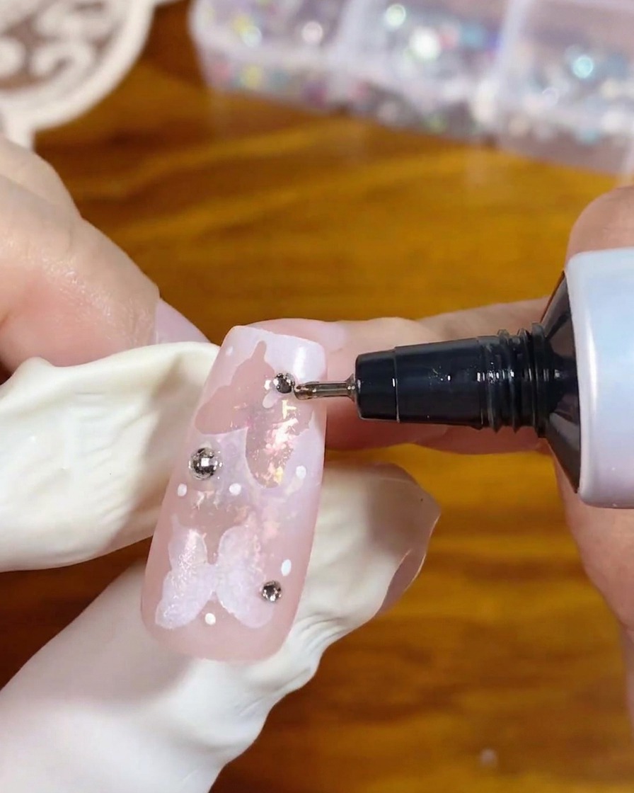 Ein Präzisionsapplikator-Tube gibt Klargel um die Basis eines silbernen Strasssteins auf einem dekorierten rosa Nagel ab, um ihn zu sichern.