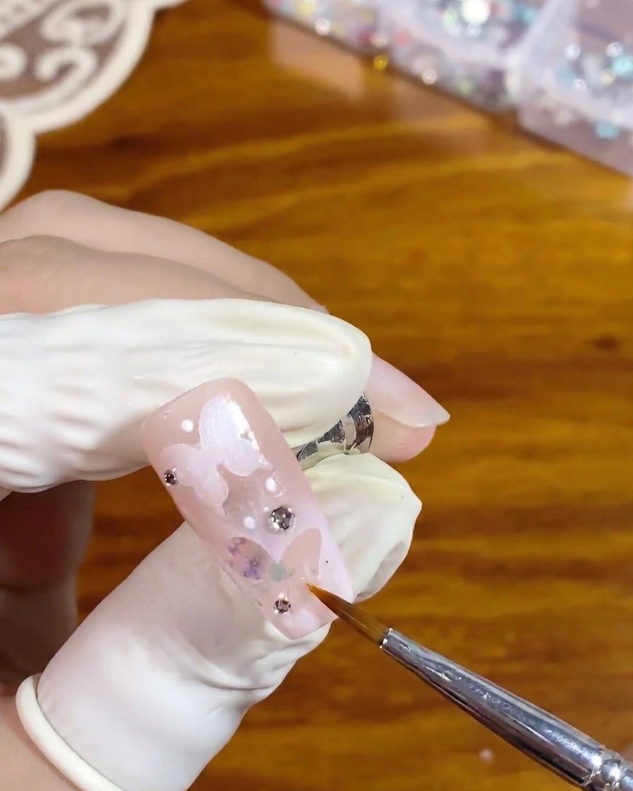 Ein feiner Liner-Pinsel trägt sorgfältig klaren, glänzenden Überlack um einen silbernen Strassstein auf einem rosa Nagel mit weißem, hohlen Schmetterlingsdesign auf.
