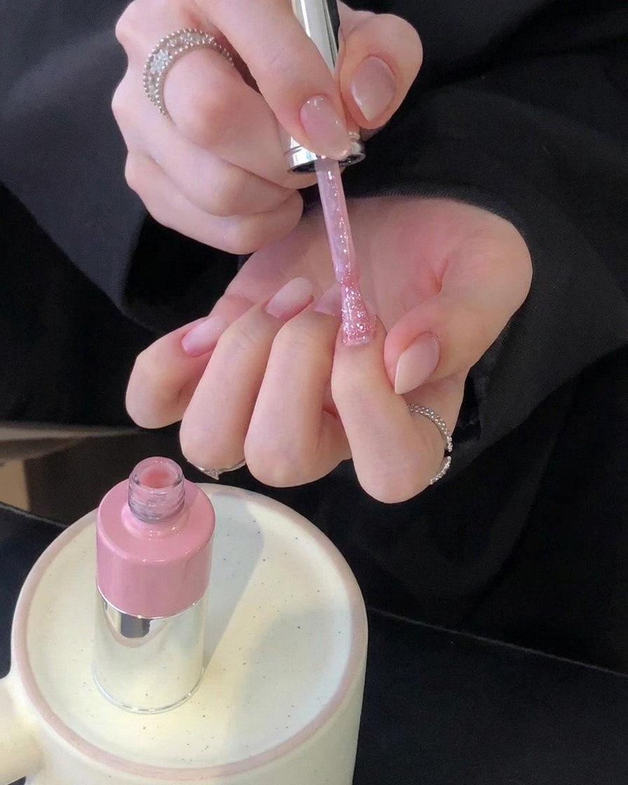 Ein Paar Hände, die einen Pinsel mit rosa Nagellack über frisch manikierten Nägeln halten, während eine rosa Flasche auf einem Keramikhalter steht.