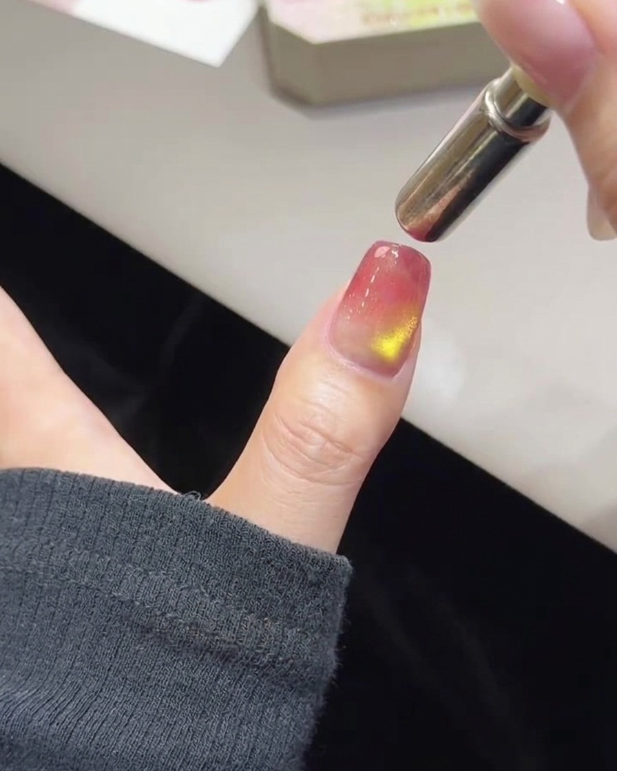 Nahaufnahme eines Daumens mit unlackiertem rosa und goldenem Lack und einem zylindrischen Magnetstab, der an der Nagelspitze schwebt.