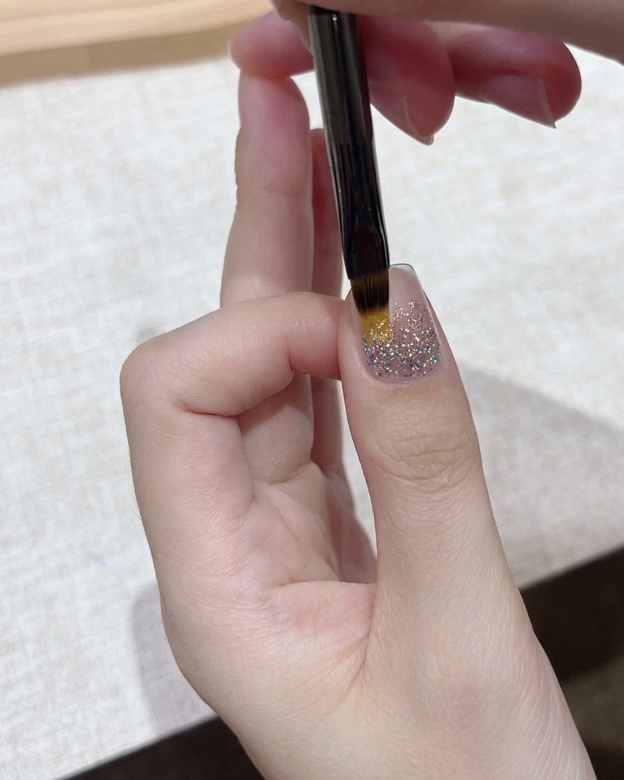 Eine Nahaufnahme eines feinen Detailpinsels, der eine Schicht klares Gel über eine funkelnde Glitzer-Nagelgrundierung aufträgt.