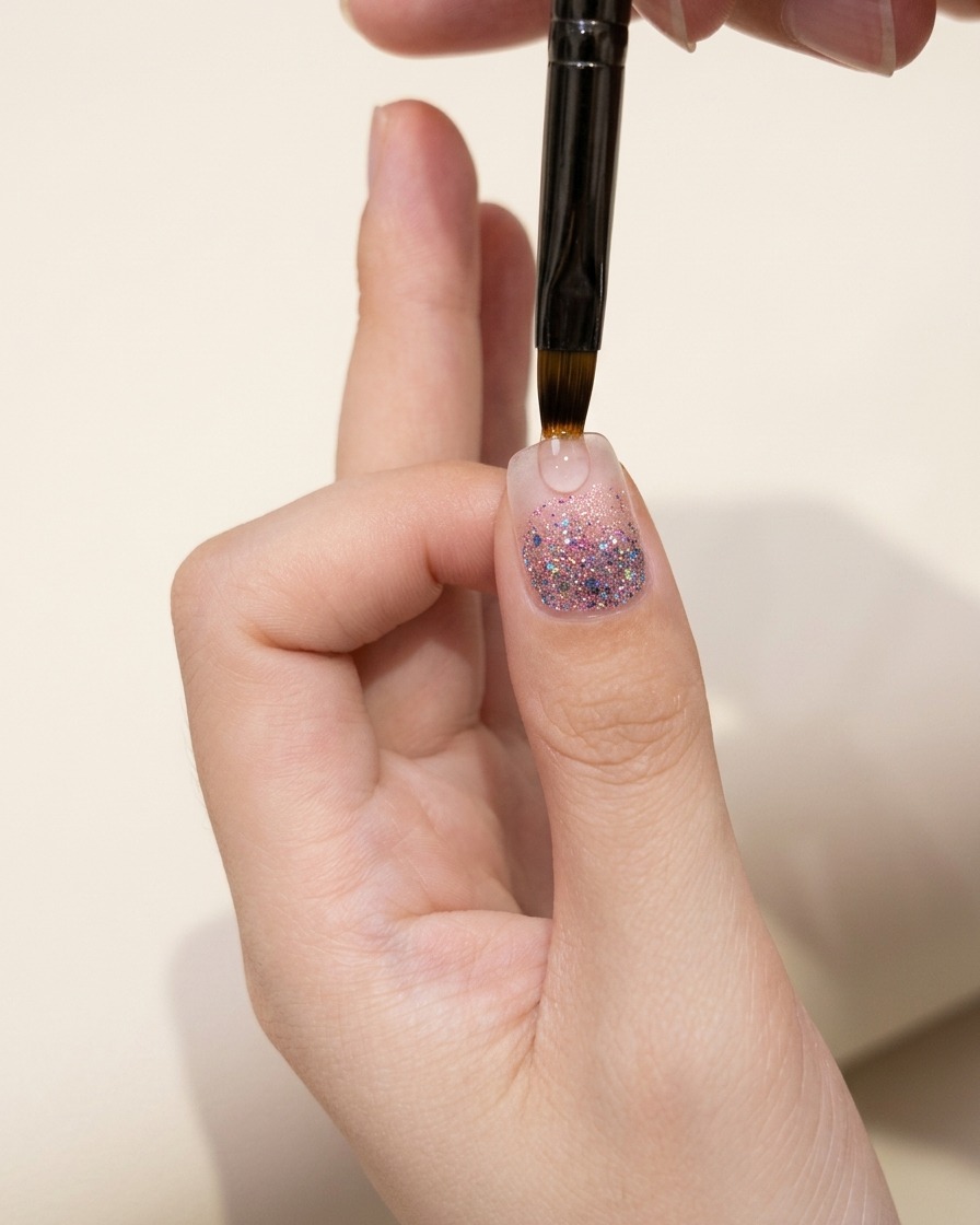 Eine Nahaufnahme eines Daumennagels mit einer Glitzer-Verlaufsschicht, die unter warmem Licht inspiziert wird.