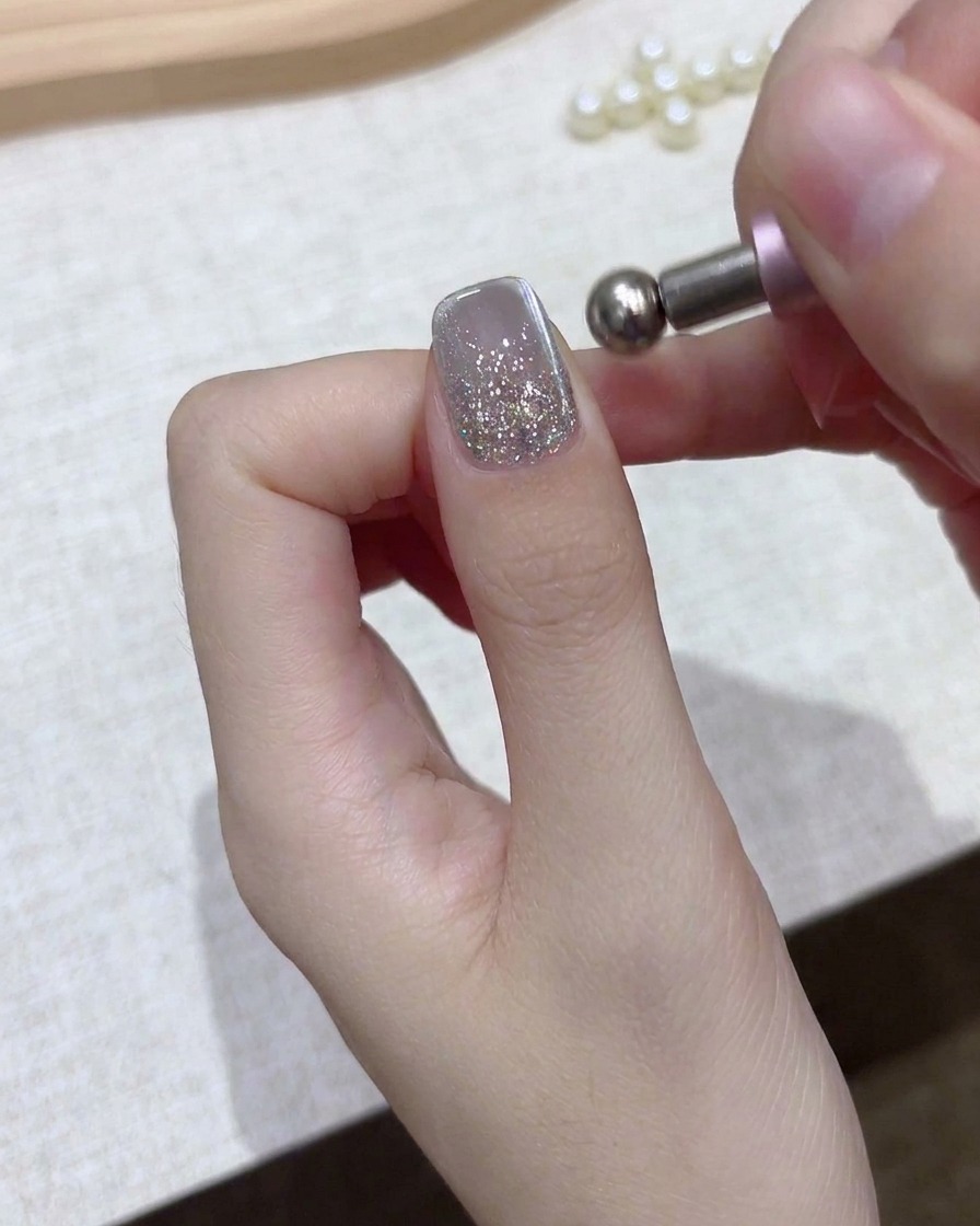 Eine Nahaufnahme einer Hand, die ein kugelförmiges Magnetwerkzeug über einem silbernen Glitzer-Gelnagel hält.