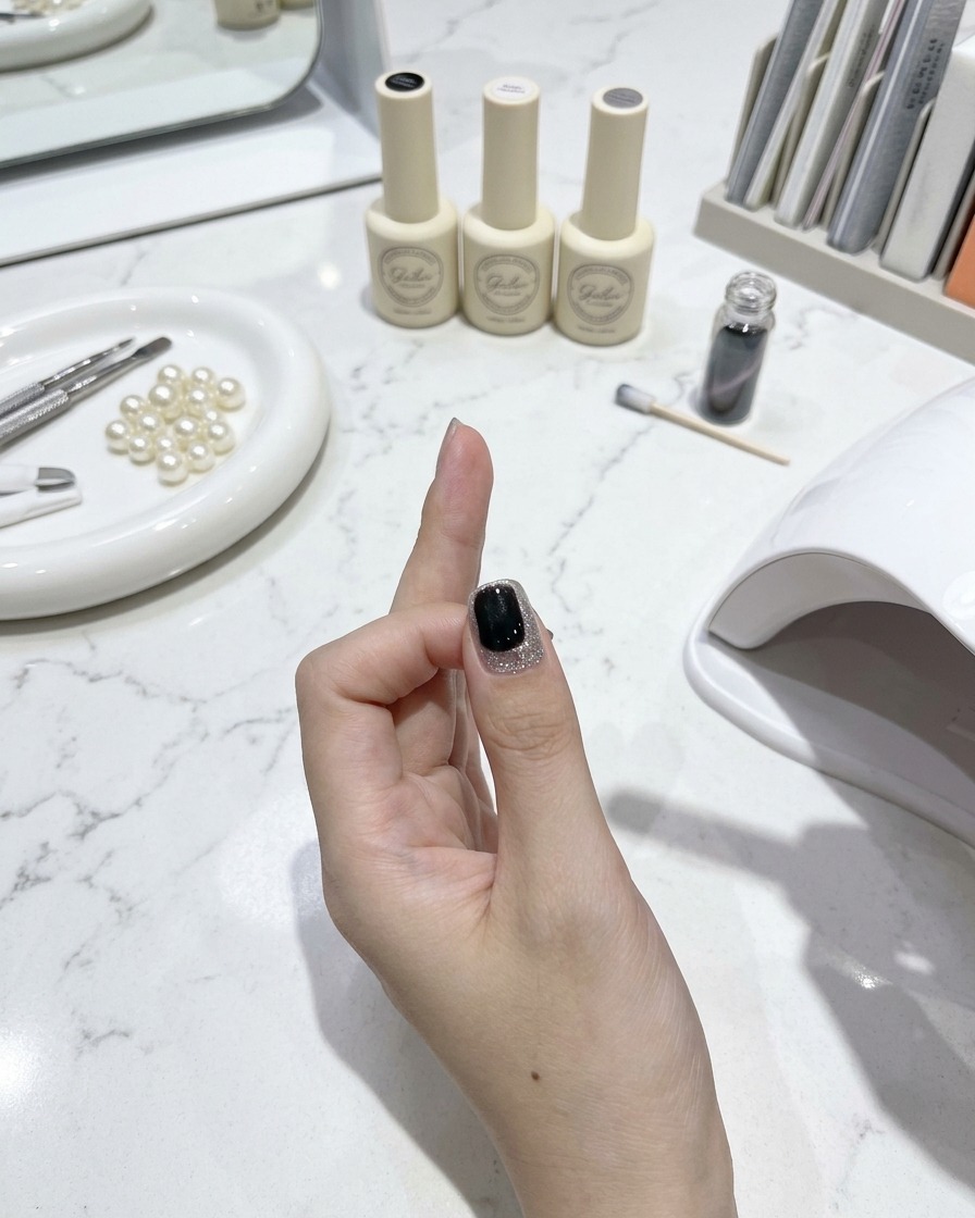 Ein Nagelpinsel trägt einen glänzenden Topcoat auf einen fertigen Cat-Eye-Nagel auf, mit Manikürewerkzeugen und Lackflaschen auf einem weißen Marmortisch im Hintergrund.