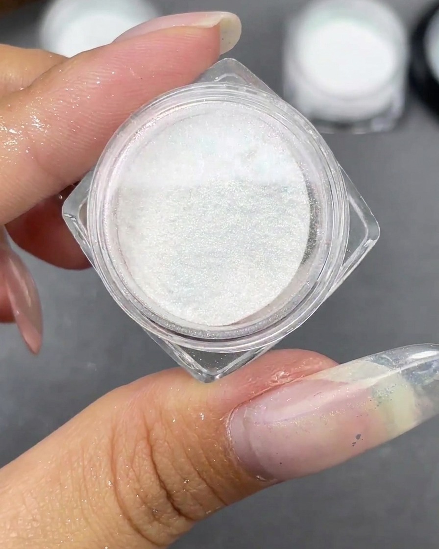 Ein kleines Glas mit weißem Moonlight-Pulver offen neben einer klaren Stiletto-Nagelspitze.