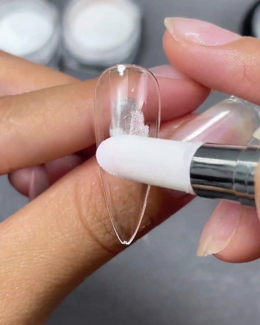 Ein Schwammapplikator tupft weißes Aurora-Pulver auf eine klare Stiletto-Nagelspitze.