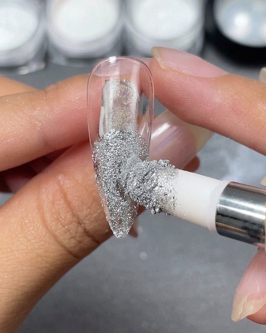 Ein Schwammapplikator gibt silbernes Chrompulver auf eine transparente Stiletto-Nagelspitze, die zwischen den Fingern gehalten wird.