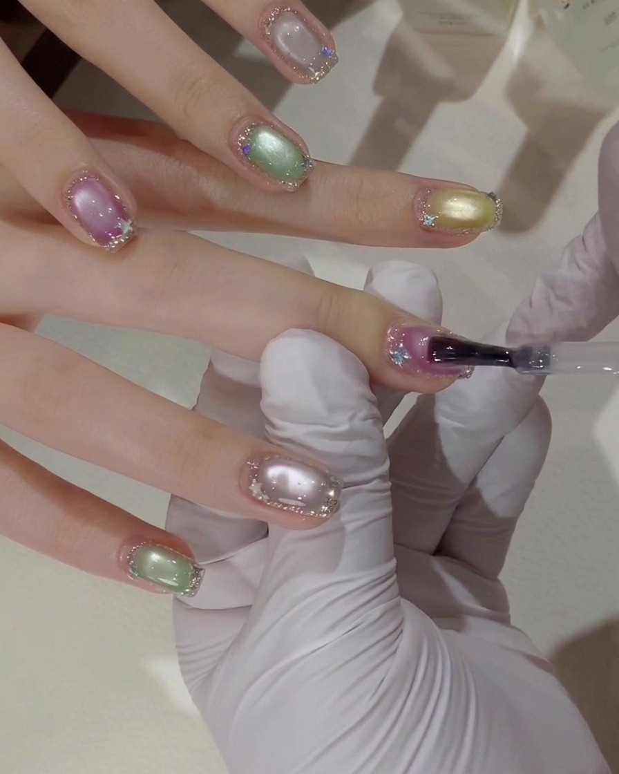 Eine Nahaufnahme einer bemalten Hand mit einem Gelpinsel, der eine Schicht klaren Topcoat über einem rosa-lila Cat-Eye-Nagel mit silbernen Glitzer-Rändern aufträgt.