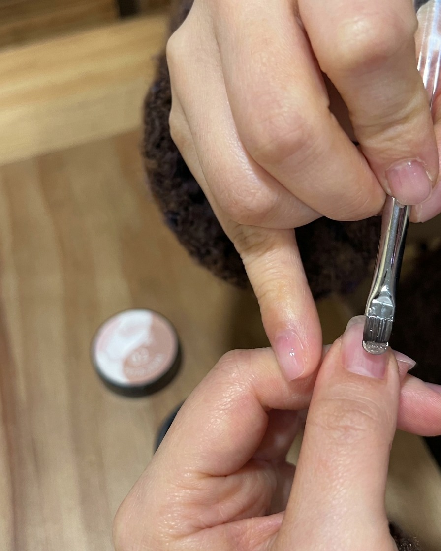 Eine Nahaufnahme eines Gelpinsels, der schimmernden Nude-Gold-Lack auf einen Naturnagel aufträgt.