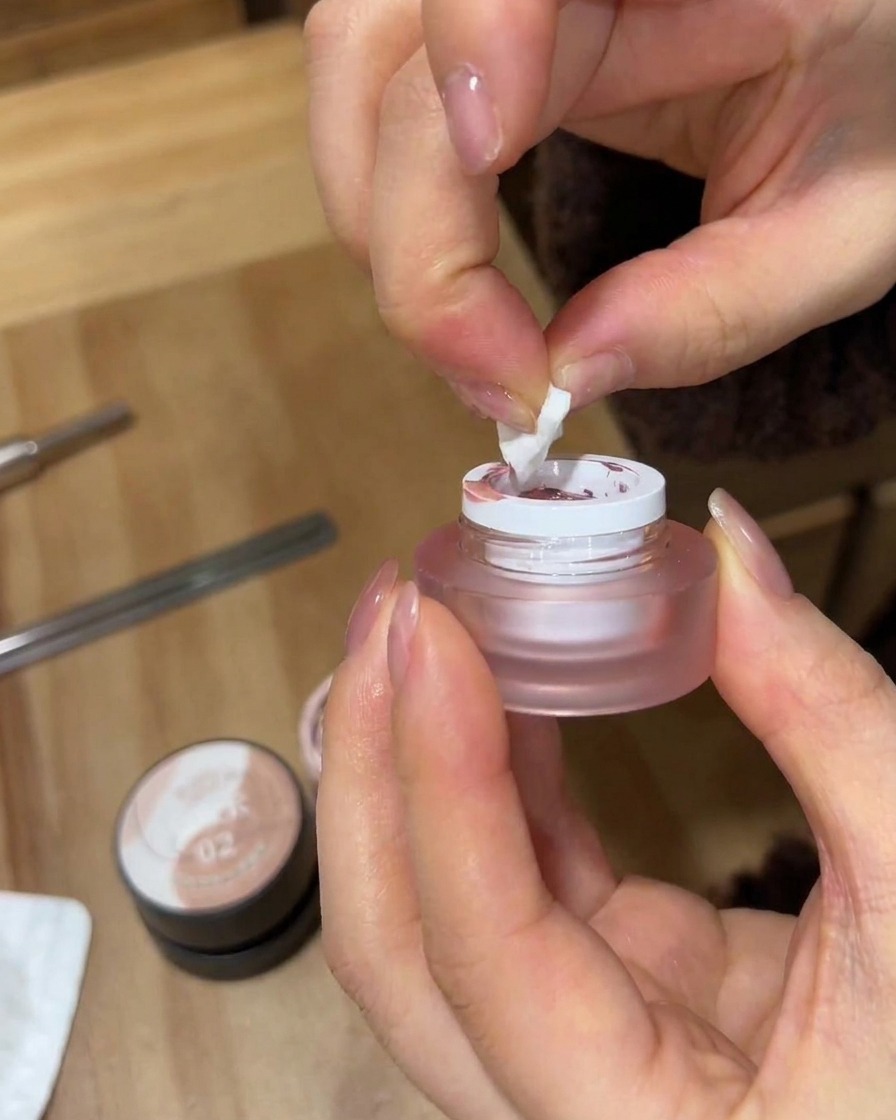 Eine Nahaufnahme von Händen, die ein zerrissenes Stück weißen Make-up-Schwamms verwenden, um rosa Gel-Nagellack aus einem kleinen mattierten Tiegel aufzunehmen.