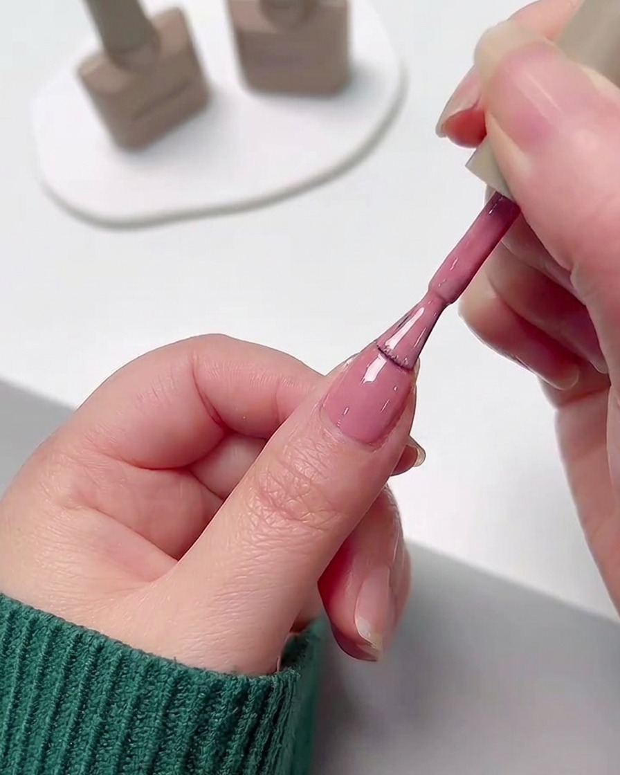 Eine Hand trägt mit dem Pinsel der Lackflasche eine glatte Schicht rosa Gel-Nagellack auf einen Fingernagel auf.