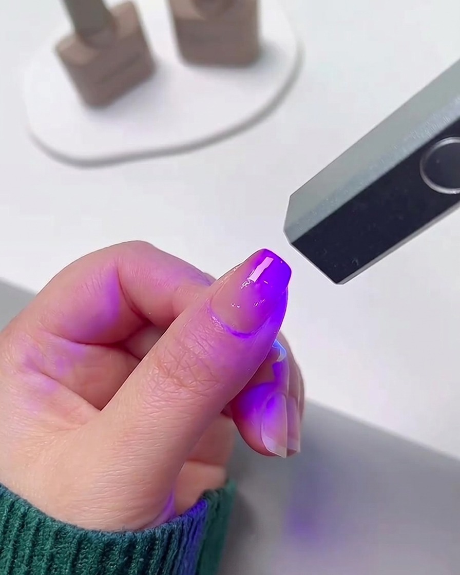 Ein Finger befindet sich unter einer kleinen, tragbaren UV- und LED-Nagellampe, die ein helles violettes Licht ausstrahlt, um den Gel-Nagellack auszuhärten.