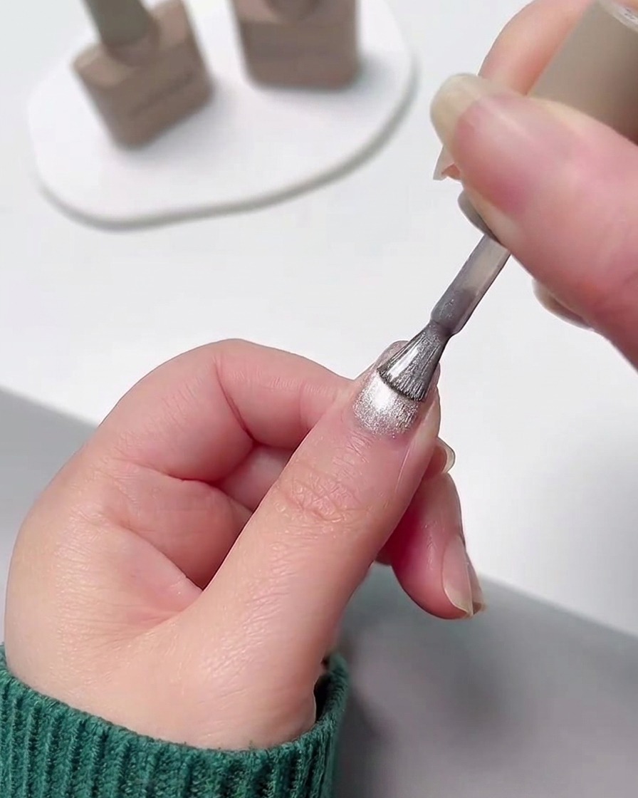 Eine Hand trägt eine Schicht schimmernden silbernen Cat Eye Gel-Nagellacks über einem ausgehärteten rosa Farbverlaufsnagel auf.