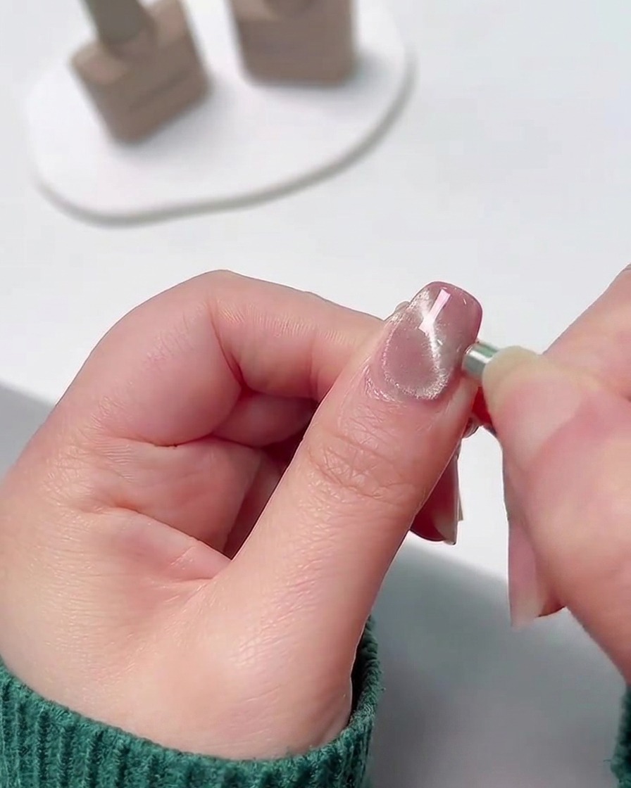 Eine Nahaufnahme eines kleinen zylindrischen Magneten, der an die Seitenkante eines feuchten, schimmernden Gelnagels gehalten wird, um die Metallpartikel in eine konzentrierte Linie zu ziehen.