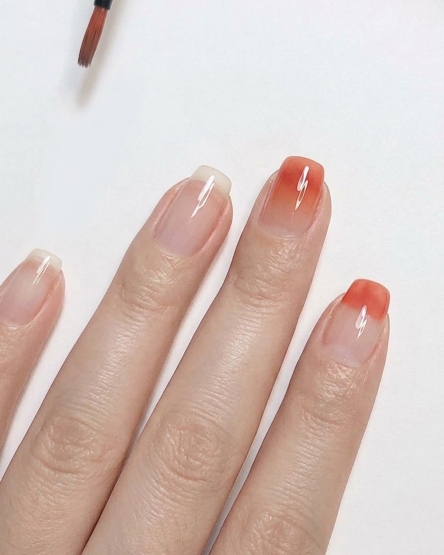 Ein feiner Detailpinsel verblendet pfirsichrosa Gel-Nagellack nach oben auf einem klaren Nagel, um einen sanften Ombré-Verlaufseffekt zu erzeugen.