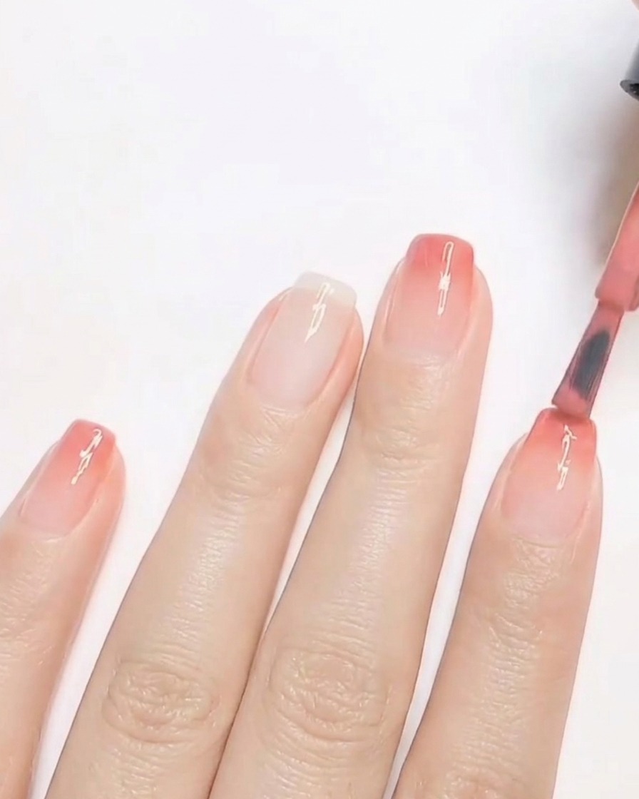Ein Nagelpinsel trägt eine zweite Schicht warmen pfirsichtönten Gel-Nagellacks auf die Spitze eines Fingernagels auf, um einen Ombré-Verlauf aufzubauen.