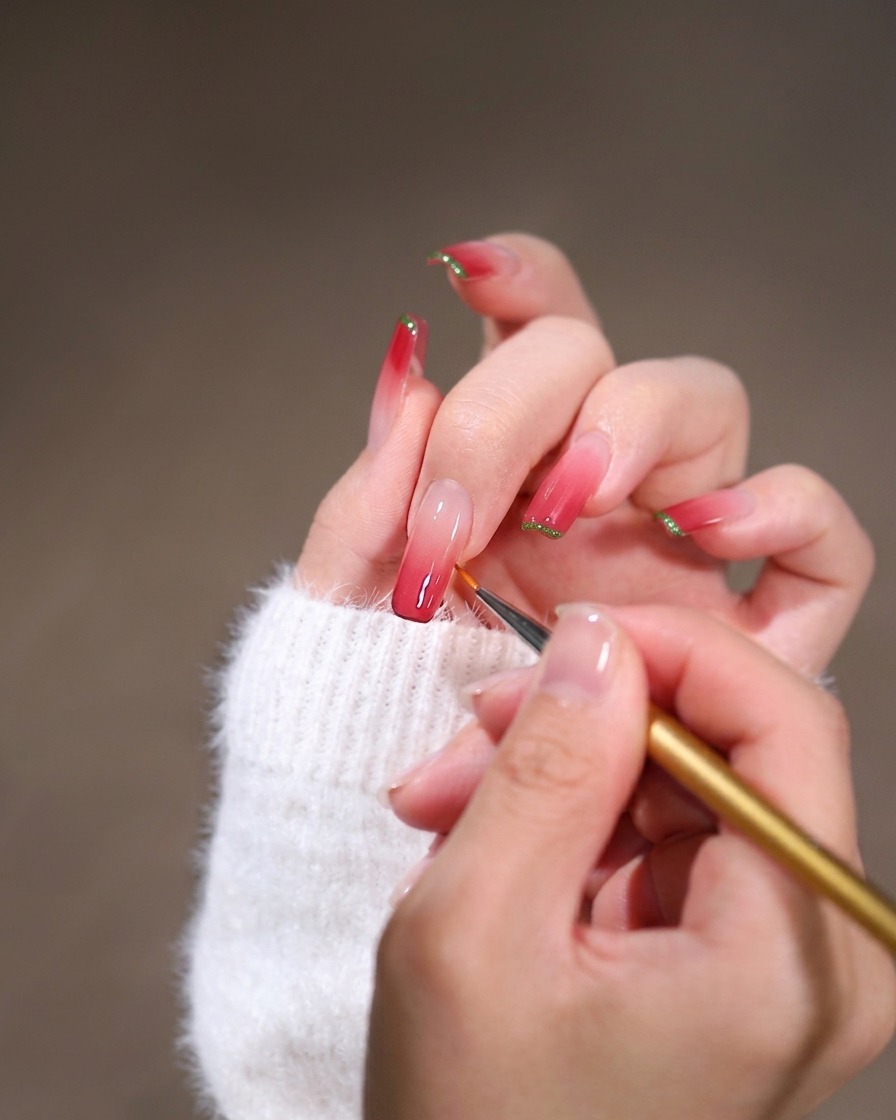 Eine Nahaufnahme eines feinen Liner-Pinsels verblendet rosa Gel-Lack entlang der Nagelhautkante eines langen quadratischen Nagels.