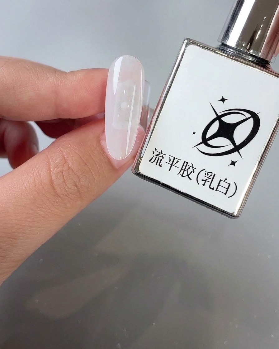 Ein Pinsel trägt milchig-weißen Selbstverlaufsgel auf eine mandelförmige Nageloberfläche neben der Produktflasche auf.