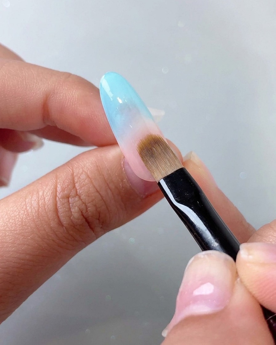 Ein Tupfpinsel verblendet einen blauen Farbverlaufseffekt auf dem oberen, rötlichen Teil des Nagels.