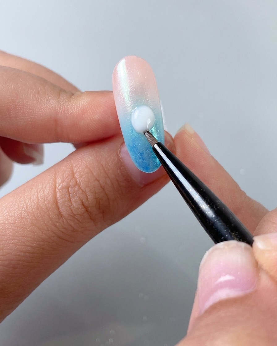 Ein feiner Nail-Art-Pinsel trägt einen Tropfen milchig-weißen Selbstverlaufsgels diagonal über einen Gradientennagel auf.