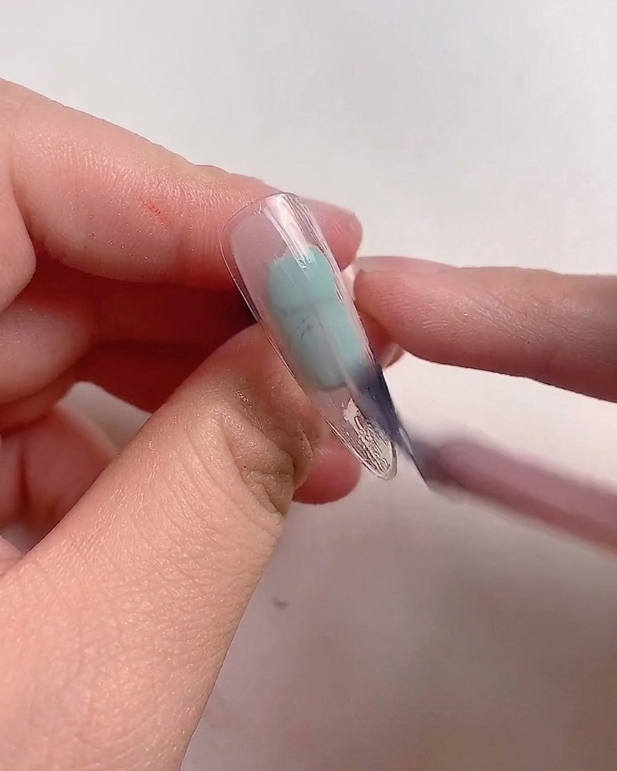 Ein Pinsel trägt mauve-pinken Gellack auf einen klaren Nageltip auf.