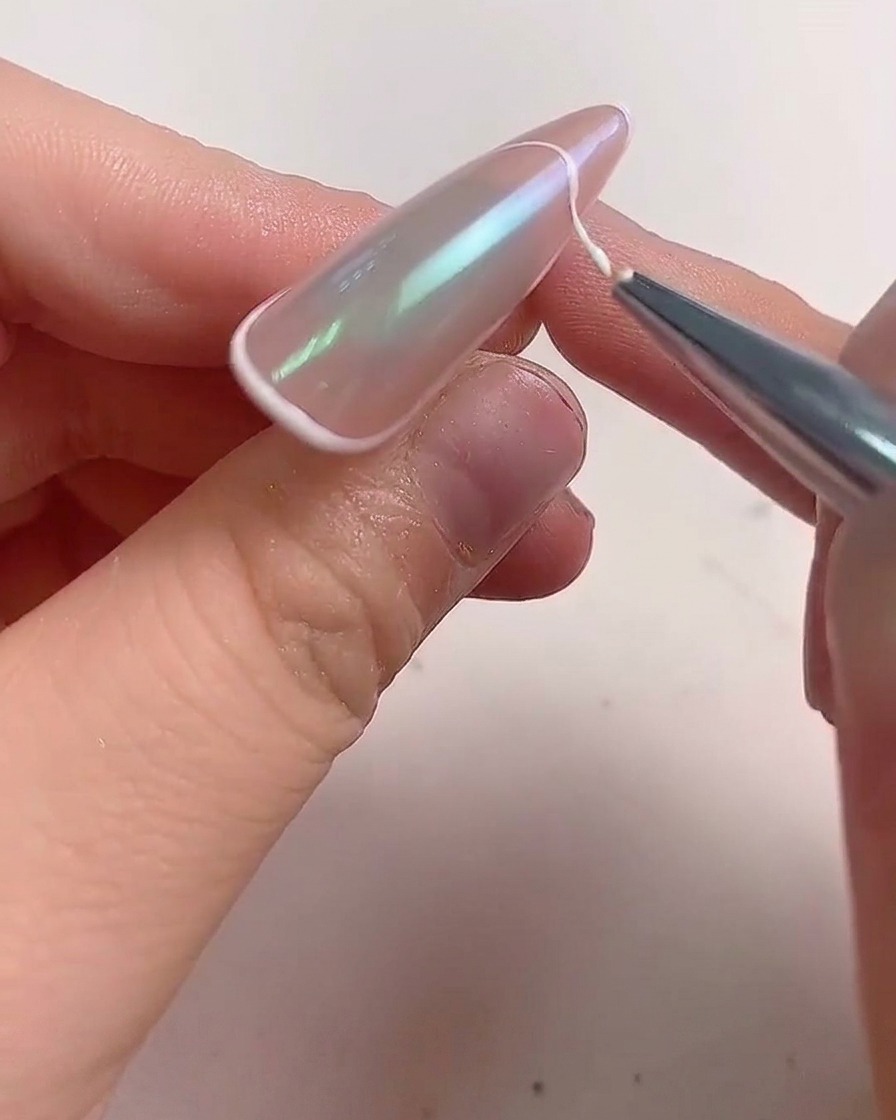 Ein feiner Liner-Pinsel zieht eine weiße Gellinie entlang der Kante eines mandelförmigen Nagels, um einen French Tip zu kreieren.