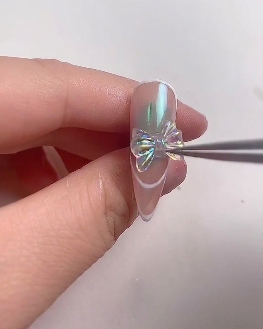 Eine Metallpinzette positioniert vorsichtig eine funkelnde, irisierende Kristallschleife in der Mitte eines mandelförmigen Nagels.