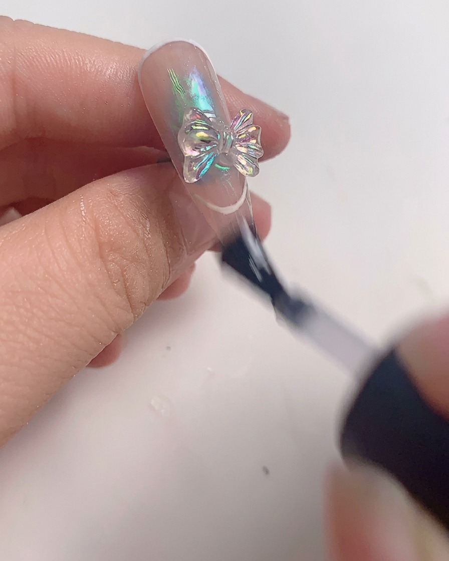 Ein Pinsel trägt eine letzte Schicht klaren Top Coat um eine Kristallschleife auf einem fertigen Nagel auf.