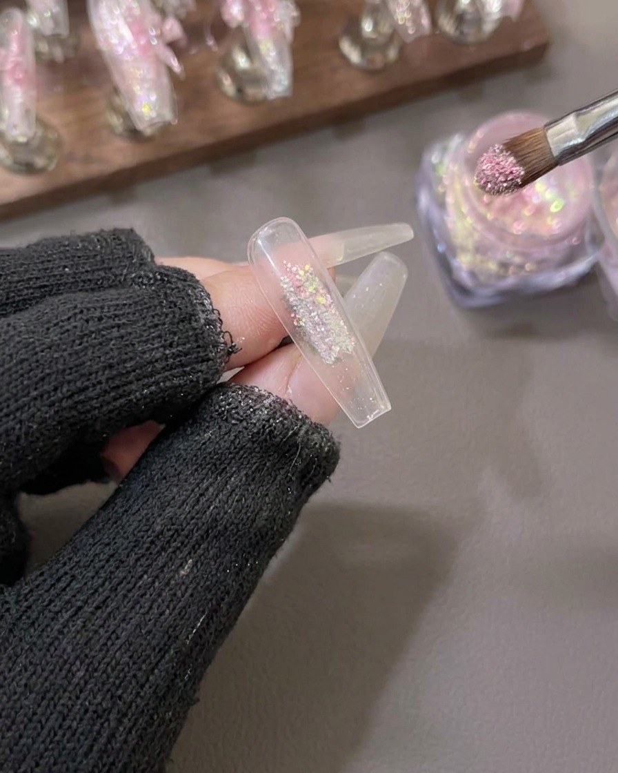 Ein Pinsel trägt funkelndes rosa und goldenes Aurora Flake Puder auf die Mitte eines klaren künstlichen Nagelkillers auf.