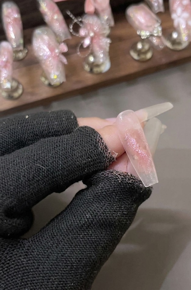 Eine behandschuhte Hand hält einen klaren Nagel, der mit einer schimmernden Schicht aus rosa Shattered Diamond Glitter Gel beschichtet ist.