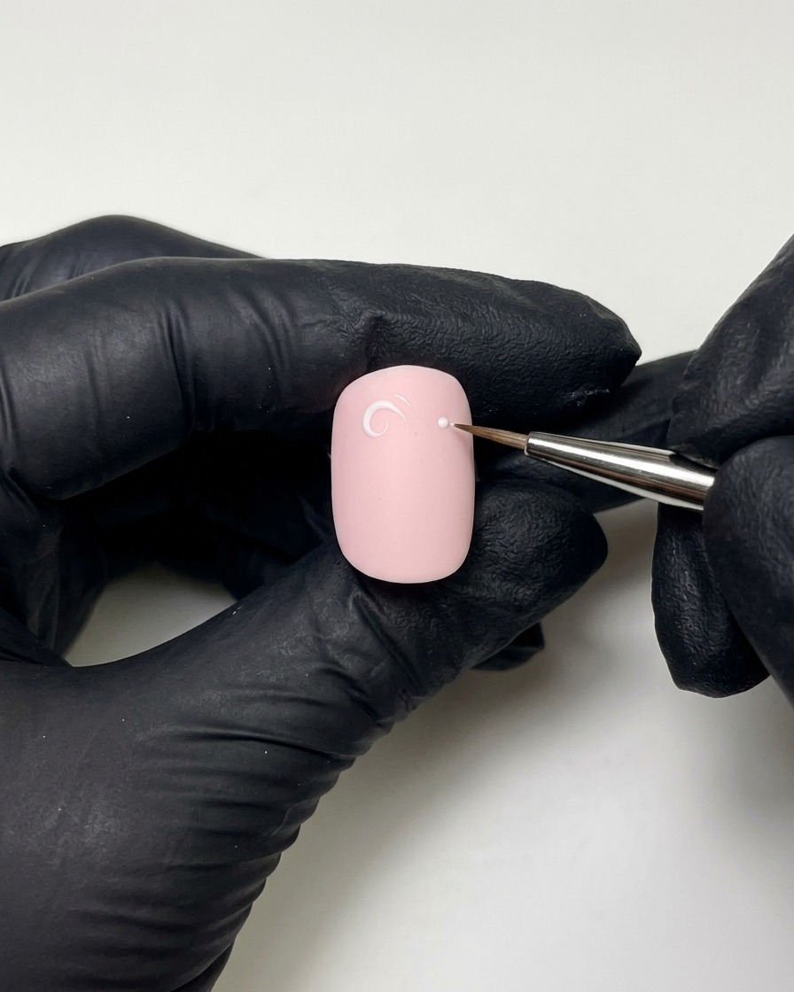 Beweumte Hände zeichnen mit einem feinen Liner-Pinsel und Aufbau-Gel eine weiß-rosa Schlaufe und einen Punkt auf einen lackierten Nagel.