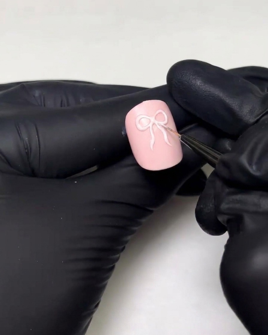 Eine be Handschuhe getragene Hand verwendet einen feinen Detailpinsel, um die Schlaufen und Bänder eines 3D-Schleifendesigns auf einen rosa Nagel zu zeichnen.
