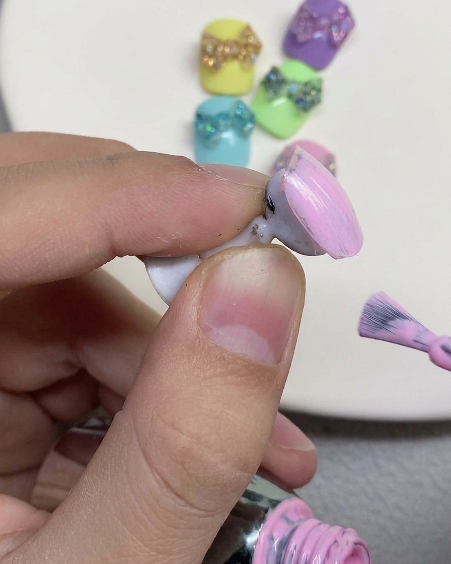 Ein Nagelpinsel trägt ein zartes, pastellrosa Farbgel auf die Oberfläche eines künstlichen Nagels auf einem Halter auf.