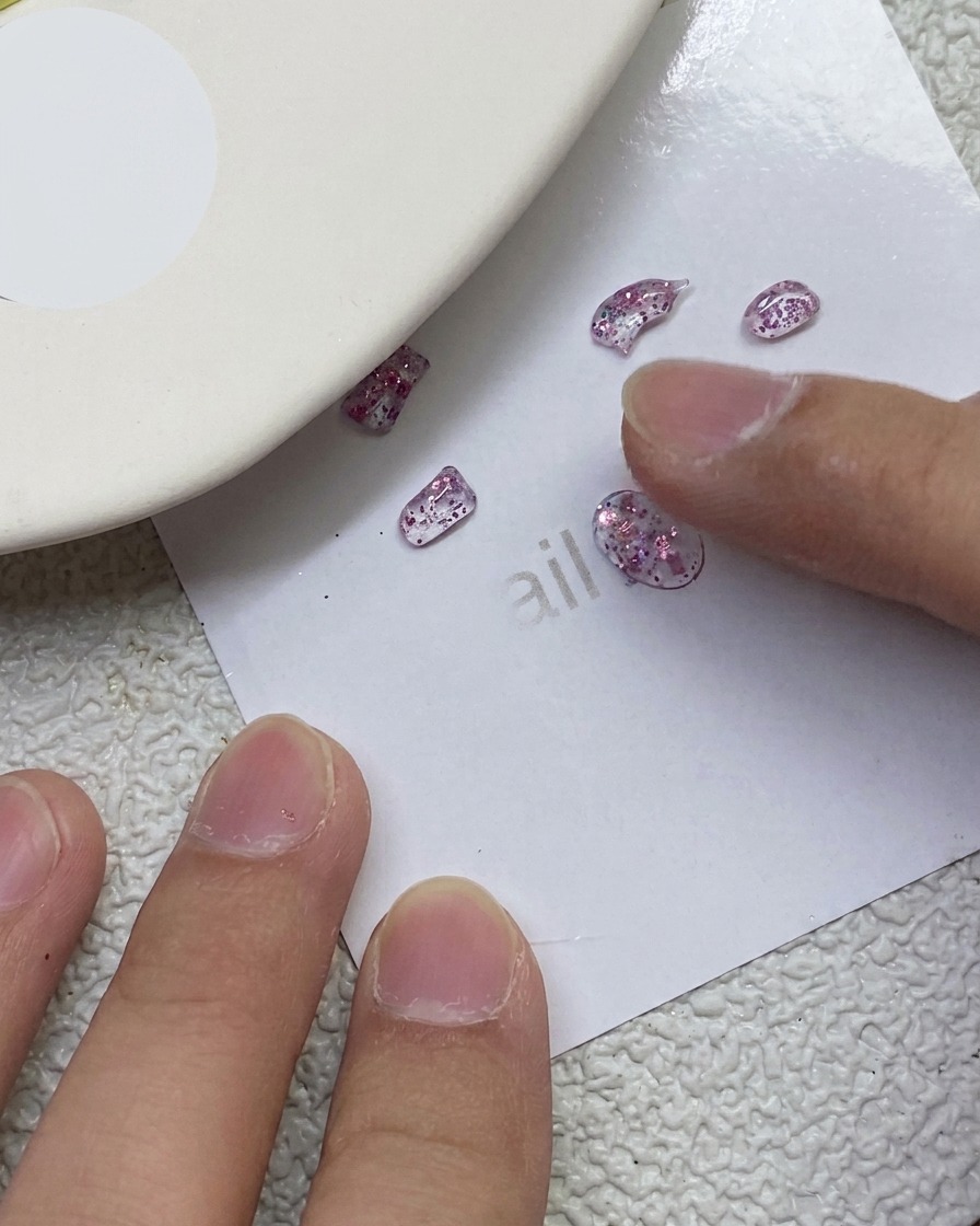 Ein Finger drückt und formt kleine rosafarbene Glitzer-PVC-Gelteile zu Schleifenformen auf einem weißen Blatt Papier.