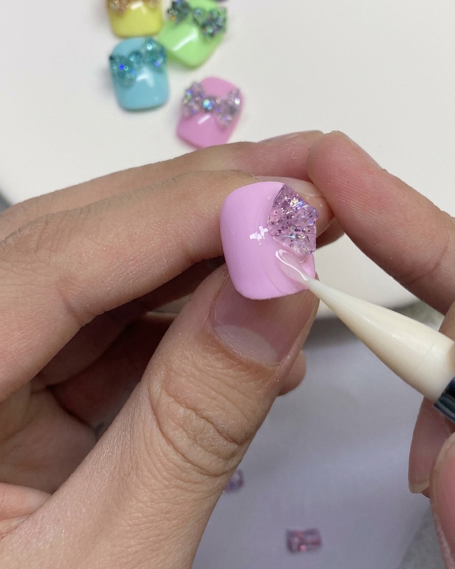 Nahaufnahme eines Silikonwerkzeugs, das Glitzergelstücke zu den äußeren Schlaufen einer Schmetterlingsschleife auf einem rosa Nagel formt.